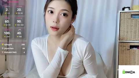 Media: Video of an East Asian woman with fair skin, dark hair tied back, wearing a white long-sleeve top, sitting on a bed with a white curtain background.