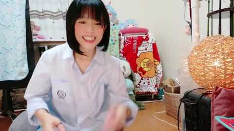 Media: Video of a smiling Asian woman with black bobbed hair, wearing a light blue button-up shirt, sitting on the floor in a cluttered, cozy room with colorful decor, including a red quilt with a bear pattern, a woven lampshade, and a wicker basket.