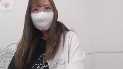 Media: Video of an East Asian woman with long brown hair, wearing a white surgical mask and white long-sleeve shirt with a black graphic design. She stands against a plain, light-colored background.