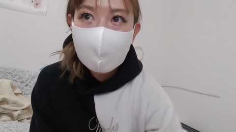 Media: Video of an Asian woman with light skin, brown hair, wearing a white surgical mask, black and white hoodie, and gray bedsheet background.