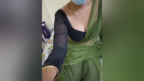 Media: Video of a woman wearing a green salwar kameez with a black, sheer top, a blue mask, and a gold bracelet, taken from an elevated angle.