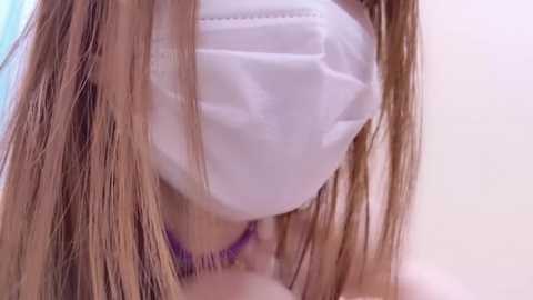 Video of a person with long, light brown hair wearing a white face mask, visible from the shoulders down. The background is a blurred, soft-focus, pastel-colored room.