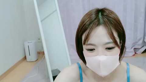 Media: Video of an East Asian woman with short brown hair, wearing a blue tank top and pink face mask, sitting on a wooden floor in a sparsely furnished room with a white curtain and a small white fan.