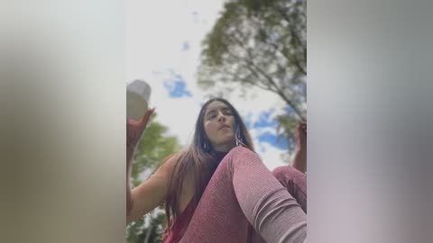 Media: A young woman with long brown hair, wearing a pink sweater and leggings, stands against a cloudy sky with green trees in the background, holding a white object.