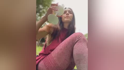 Media: Video of a young woman with long brown hair, wearing a maroon sweater and matching knit leggings, sitting on the grass, drinking from a white cup, in a park with greenery in the background.