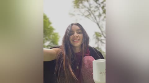 Media: Video of a young woman with long dark hair, wearing a maroon top, smiling outdoors, holding a microphone and a glass, with blurred green trees in the background.