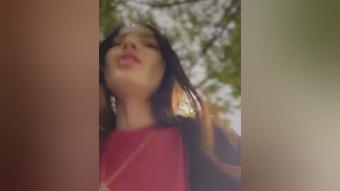 Media: A blurred video of a young woman with long, dark hair and fair skin, wearing a red top, captured from a low angle, with green foliage and sunlight in the background.