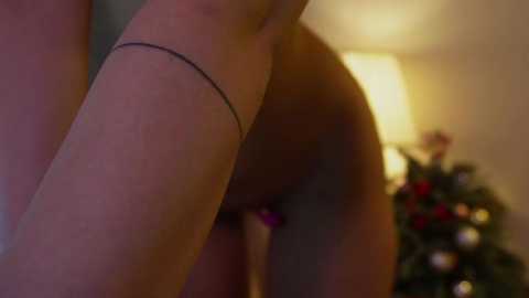 Media: Video of a close-up view of a nude woman's lower torso, showing her shaved pubic area. She has a thin, black ankle bracelet. Background features a blurred Christmas tree with colorful ornaments.