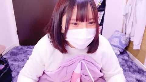 Media: Video of an East Asian woman with short dark hair and a white face mask, wearing a white long-sleeve shirt and a light pink sweater, lying on a purple blanket in a room with white walls and a wooden floor.