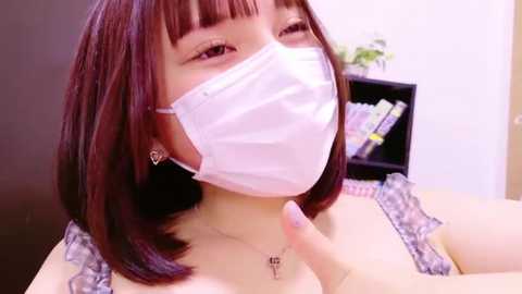 Media: Video of an Asian woman with straight brown hair, wearing a white surgical mask, a grey lace bra, and a necklace, winking, indoors.