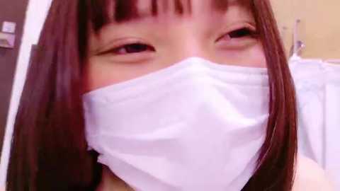 Media: Video of an East Asian woman with straight, shoulder-length dark hair, wearing a white medical mask, smiling, with a blurred background of a bathroom with a shower curtain.