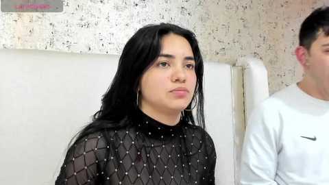 Media: Video of a young woman with long black hair, wearing a black, see-through top with diamond patterns, standing in a hospital room with a white wall and a man in a white shirt partially visible.