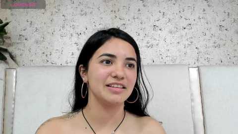 Media: Video of a young Latina woman with long black hair, wearing hoop earrings, a black necklace, and a white tank top, smiling. Background shows textured white wall and a plant.