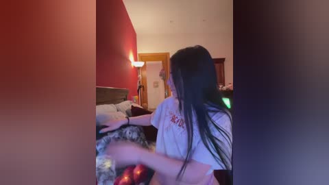 Media: Video of a woman with long black hair, wearing a white graphic t-shirt, dancing on a bed with a red blanket, in a dimly lit, red-walled room.