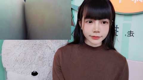 Media: Video of a young East Asian woman with straight black hair and bangs, wearing a brown turtleneck, standing in front of a colorful, cartoonish background with a large white teddy bear.