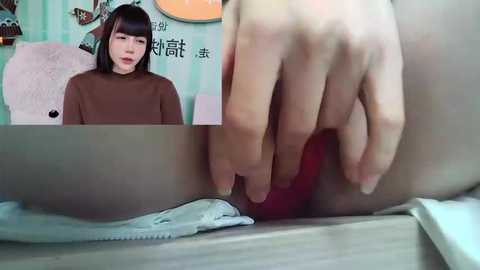 Media: Video: A young Asian woman with straight black hair, wearing a brown sweater, sits on a white chair against a green and white striped wall. A hand inserts a red object into her vagina.