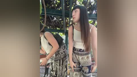 Media: Video of a young woman with long brown hair in a white tank top and patterned pants, standing in a lush, green garden with a metal structure in the background.