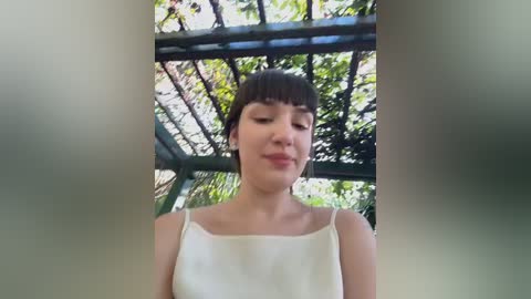 Media: A video of a young woman with short dark hair and fair skin, wearing a white tank top, standing in a sunlit garden under a pergola with green leaves and beams.