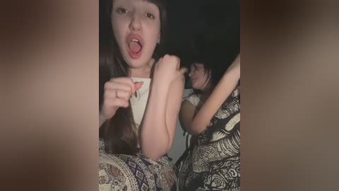 Media: Video of a young Asian girl with long brown hair, sticking out her tongue, wearing a white shirt, surrounded by patterned blankets, and another Asian girl in the background.