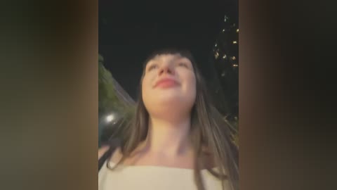 Media: A video of a young woman with long brown hair, wearing a white top, smiling upward at the night sky, surrounded by blurred greenery and lights.