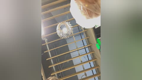 Media: Video of a modern indoor space with a chandelier hanging from a high ceiling, featuring a light bulb in a white globe. The background shows a sleek, minimalist design with horizontal wooden beams and a large, rectangular window.