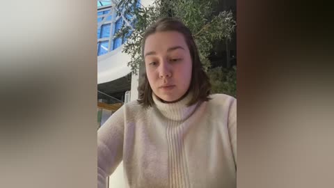 Media: Video of a young woman with shoulder-length brown hair, wearing a cream-colored turtleneck sweater, standing indoors with greenery and modern architecture in the background.