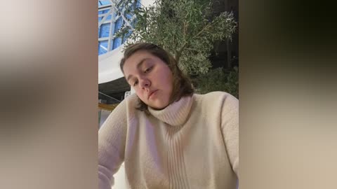 Media: A candid video captures a young woman with shoulder-length brown hair, wearing a cream-colored turtleneck sweater, leaning on a table with a relaxed, contemplative expression. The background features a modern glass structure and lush green foliage.