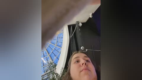 Media: Video of a young woman with light skin and brown hair looking up, taken from a low angle. She's indoors, near a glass dome ceiling and a black wall with electrical cords.