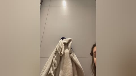 Media: Video of a woman in a beige sweater, holding a towel, standing in a bathroom with a gray tiled wall and a ceiling light.