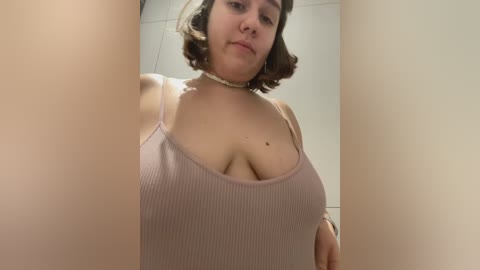 Media: Video of a curvy, light-skinned woman with shoulder-length brown hair, wearing a ribbed, pink tank top that reveals her large breasts, taken from a low angle. She has a neutral expression.