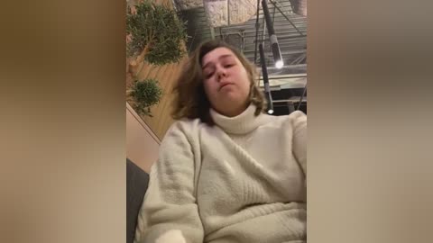 Media: A video captures a young, fair-skinned woman with shoulder-length brown hair, wearing a cream-colored, chunky-knit sweater, slouched in a chair. The background features a faux tree and a metal structure, giving a cozy, indoor feel.