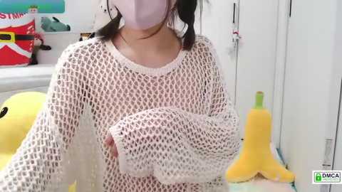 Video of a person wearing a white, fishnet crochet sweater, a white face mask, and holding a yellow plush toy in a brightly lit room with colorful toys and stuffed animals in the background.