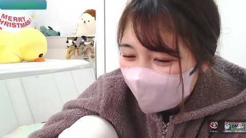 Video of a young Asian woman with medium-length black hair, wearing a pink face mask, brown fuzzy jacket, and white shirt, sitting in a brightly lit room with a \"Merry Christmas\" sign, plush toys, and a yellow stuffed duck.