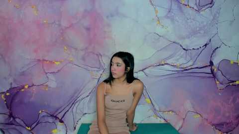 Media: Video of a young Asian woman with straight black hair, wearing a beige Gucci tank top, seated against a colorful, abstract, marble-patterned backdrop.