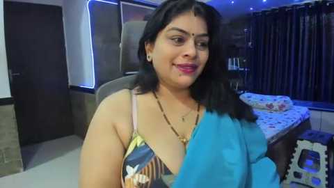 Media: Video of a smiling, dark-skinned Indian woman with long black hair, wearing a blue sari and a colorful blouse, sitting on a bed in a dimly-lit room with blue curtains.