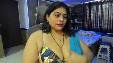 Media: A video shows a South Asian woman with medium skin tone and dark hair, wearing a colorful bra with a blue top, sitting in a cozy bedroom with a bed, curtains, and a chair.