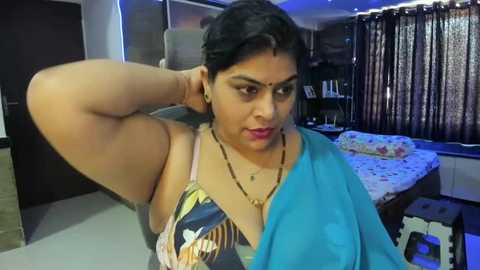 Media: Video of a South Asian woman with medium skin tone, wearing a blue saree, showing her armpit. Background shows a dimly lit bedroom with a bed, curtains, and a dark door.