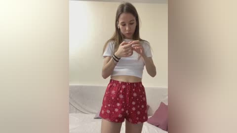 Media: A video of a young woman with light skin and long brown hair, wearing a white crop top and red pajama shorts, playing with her phone in a simple, beige room with a bed and pillows.