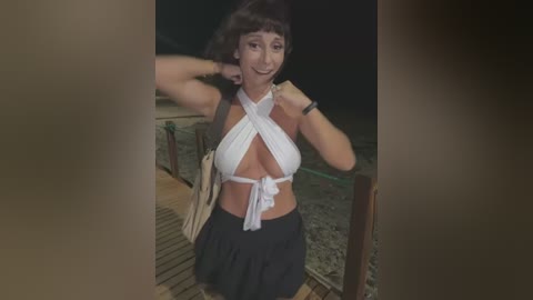 Media: A video of a smiling, light-skinned woman with short brown hair, wearing a white halter top and black skirt, standing on a wooden dock at night.