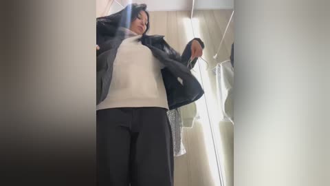 Media: Video of a woman in a white sweater and black pants, adjusting a black jacket in a bright, modern room with white walls and a window with sheer curtains.