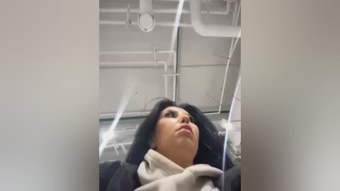 Media: Video of a woman with long black hair and fair skin, wearing a beige scarf and dark jacket, lying on her back in an industrial, white-tiled room with exposed pipes and vents on the ceiling.