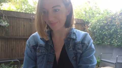 Media: Video of a smiling woman with straight, shoulder-length brown hair, wearing a denim jacket over a black top, standing in a garden with a wooden fence and greenery in the background.