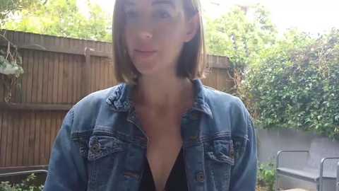 Media: Video of a young Caucasian woman with straight, shoulder-length brown hair, wearing a blue denim jacket and a black top, standing in a sunlit garden with a wooden fence and greenery in the background.