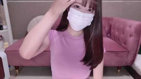 Media: A video of an East Asian woman with straight, long black hair, wearing a white surgical mask, light pink top, and sitting on a pink, tufted velvet couch. Background includes a beige wall and white furniture.