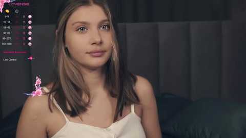 Media: Video of a young Caucasian woman with straight, shoulder-length brown hair, wearing a white spaghetti-strap top, against a dark background. She has a neutral expression.