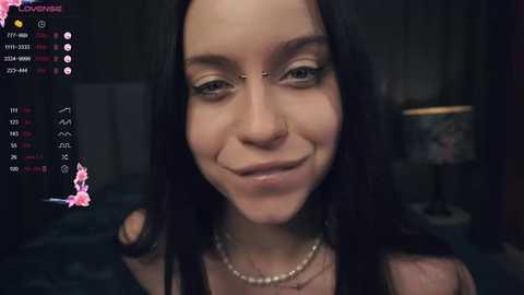 Media: Video of a smiling, pale-skinned woman with long black hair, nose piercing, and multiple facial tattoos, wearing a pearl necklace, indoors.