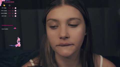 Media: Video of a young girl with long, straight brown hair, fair skin, and closed eyes, wearing a white tank top. Background shows a dimly lit room with dark furniture.