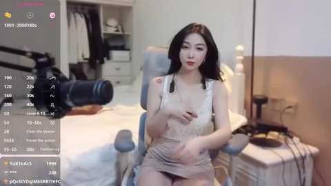 Media: A video shows an Asian woman with long black hair and light skin, wearing a sheer, white lace dress, sitting in a chair. A camera is pointed at her. The background includes a bed, wardrobe, and a lamp.