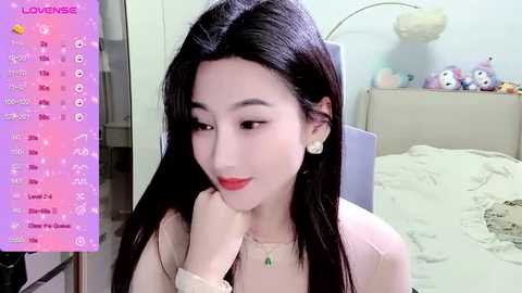 Media: A video of a young Asian woman with long black hair and light skin, wearing red lipstick and a beige top, sitting in a messy bedroom. The background features a bed with a white sheet and stuffed toys.