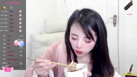 Media: A video of an East Asian woman with long black hair, wearing a pink sweater, eating noodles in a white bowl, in a bright room with a white door.